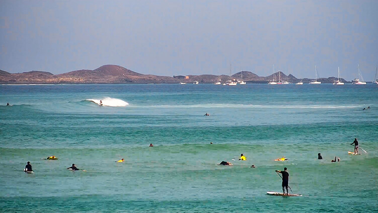 Panoramic view of Corralejo Bay live webcam at Waikiki Beach, featuring the pier for Wing Foils and Windsurfs, Rocky Point surf spot, Los Lobos Island, and the port of Corralejo with Lanzarote in the background.