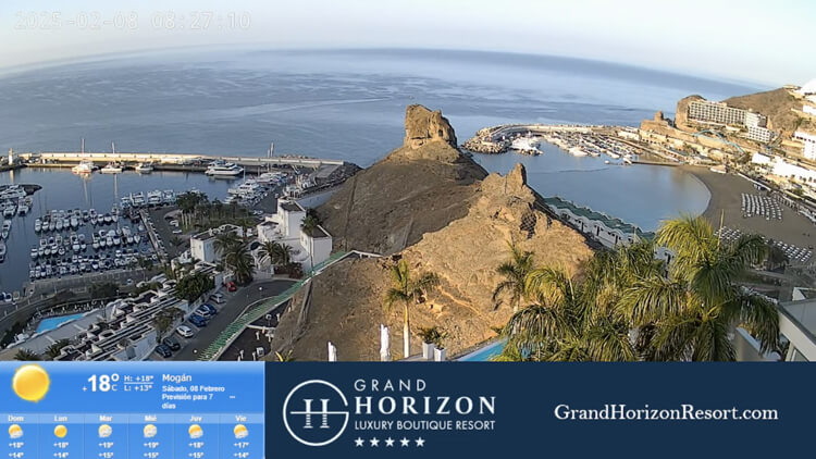 Live view of Puerto Rico Marina in Gran Canaria, showcasing the picturesque beach, calm turquoise waters, and a bustling harbor filled with yachts, restaurants, and shops.